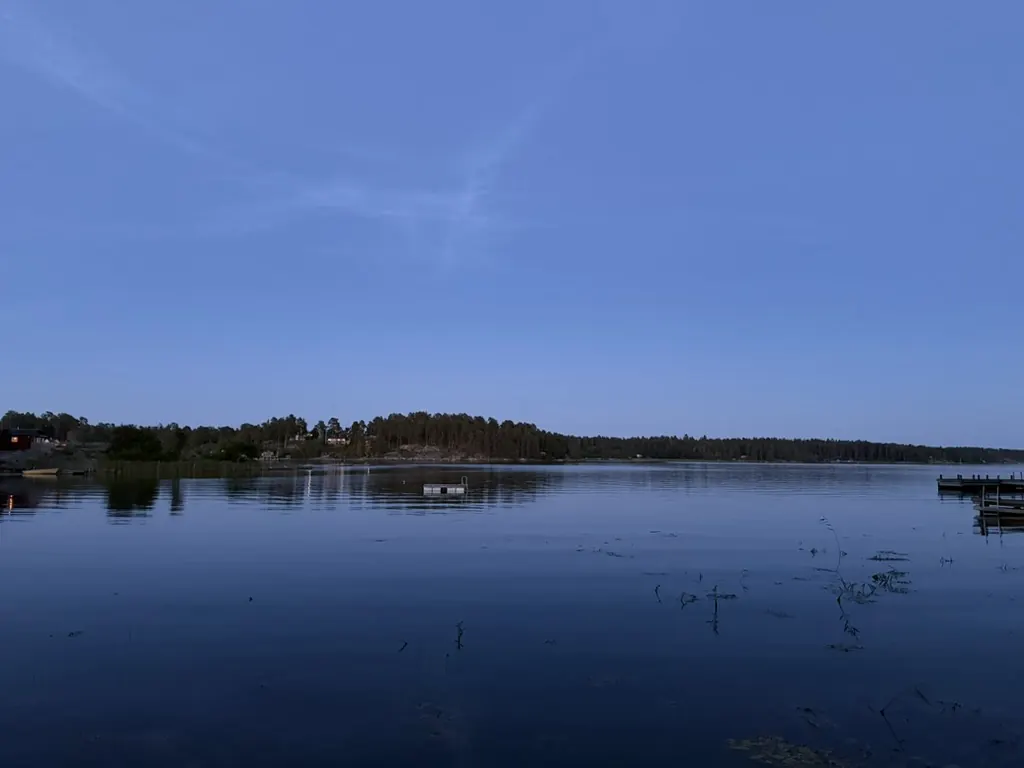 Stilla sjö i skymningen med skogsklädd strand och brygga – utsikt från stugan